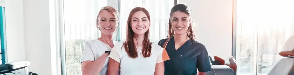 Drei Frauen stehen in einem Praxisraum, in dem mehrere Liegen stehen und lachen in die Kamera.