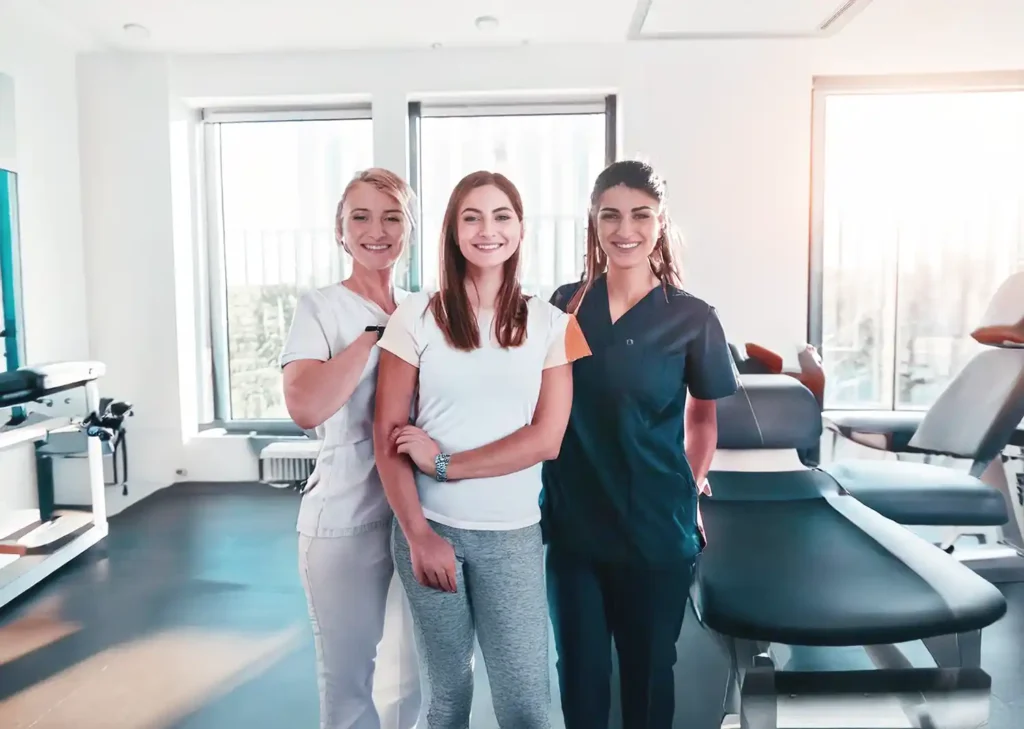 Drei Frauen stehen in einem Praxisraum, in dem mehrere Liegen stehen und lachen in die Kamera.
